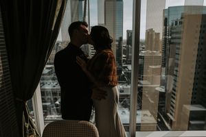 Bride and groom kissing silhouetted against city skyline through hotel window.