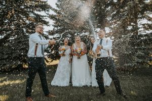 Wedding party celebrates by popping champagne bottles outdoors among tall pine trees.