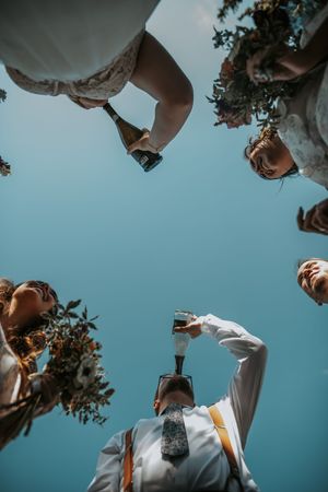 Low-angle shot of wedding party pouring champagne against a clear blue sky.
