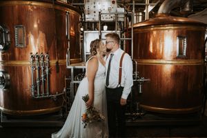 Newlyweds share a kiss in front of large copper brewery tanks.