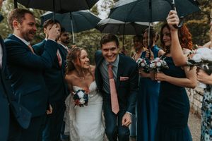 Bride and groom walk with wedding party under black umbrellas in the rain.