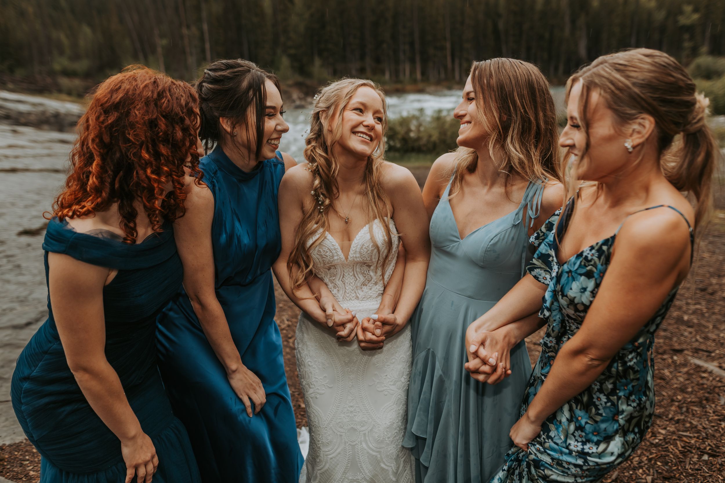 Bride laughing joyfully with bridesmaids in blue dresses near a forest riverside setting.