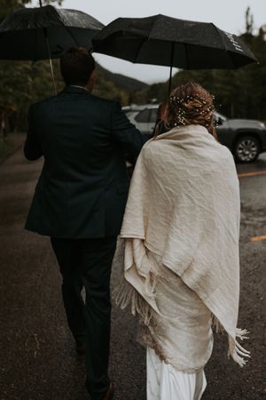 Bride wrapped in cream fringe shawl walks with groom under umbrella on rainy wedding day.