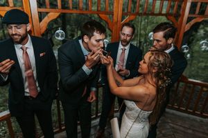 Bride and groomsmen playfully blowing bubbles together on a wooden gazebo porch.