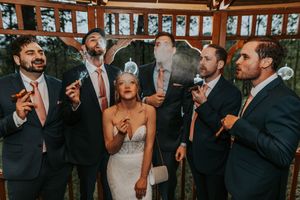 Bride and groomsmen in navy suits joyfully blowing bubbles in a rustic wooden gazebo.