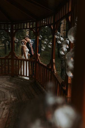 Bride and groom share a romantic kiss on a wooden gazebo porch surrounded by bubbles.