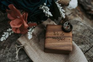 Wedding rings displayed on an engraved wooden box reading We Do beside colorful florals.