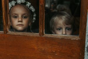 Two young children with blue eyes peek through a rustic wooden door with glass panes.
