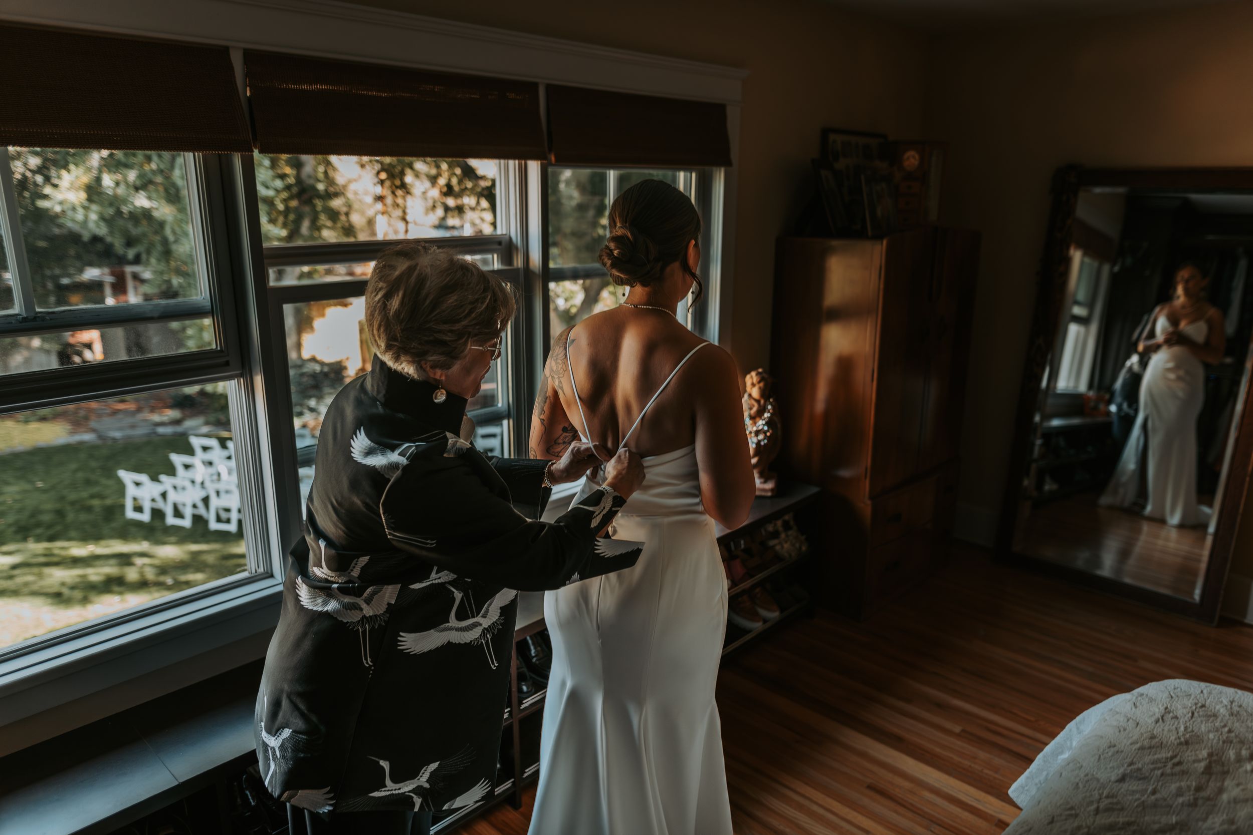 Woman helping a bride fasten her wedding dress near a bright window indoors.