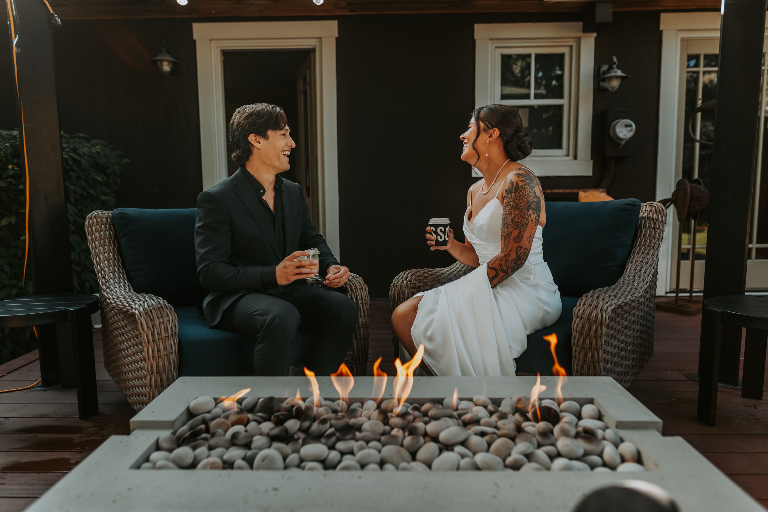 Bride and groom sharing drinks by a modern fire pit on a cozy outdoor patio at dusk.