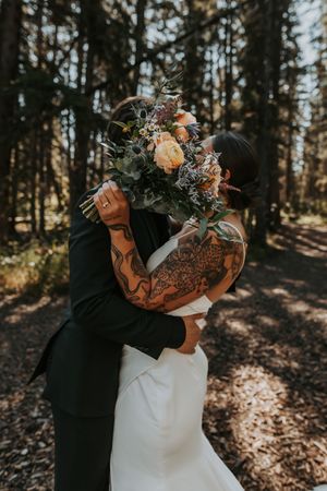 Bride and groom embracing in a forest, her tattooed arms holding a lush floral bouquet.