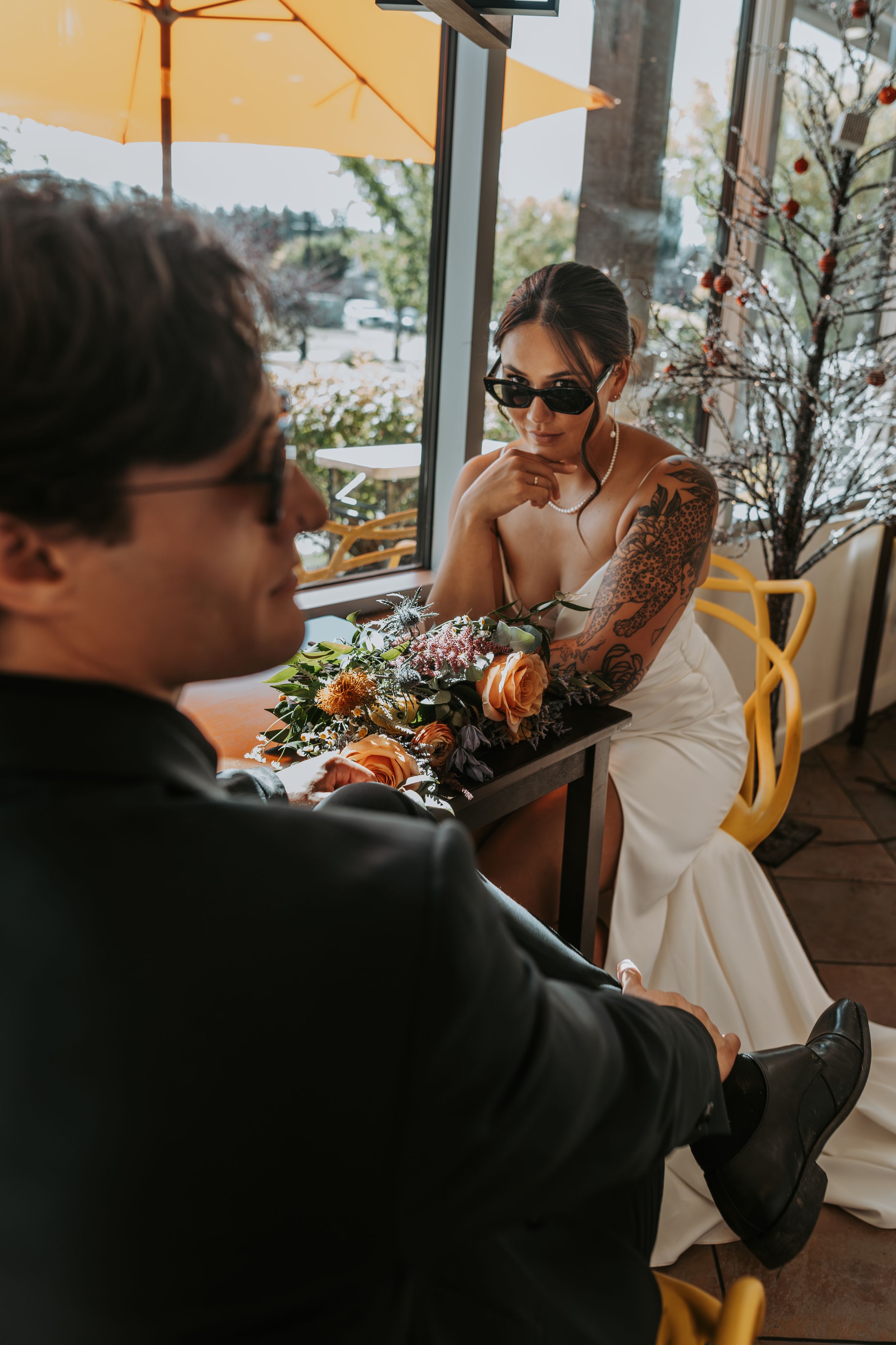 Tattooed bride in sunglasses sits with bouquet across from groom at a bright cafe window.