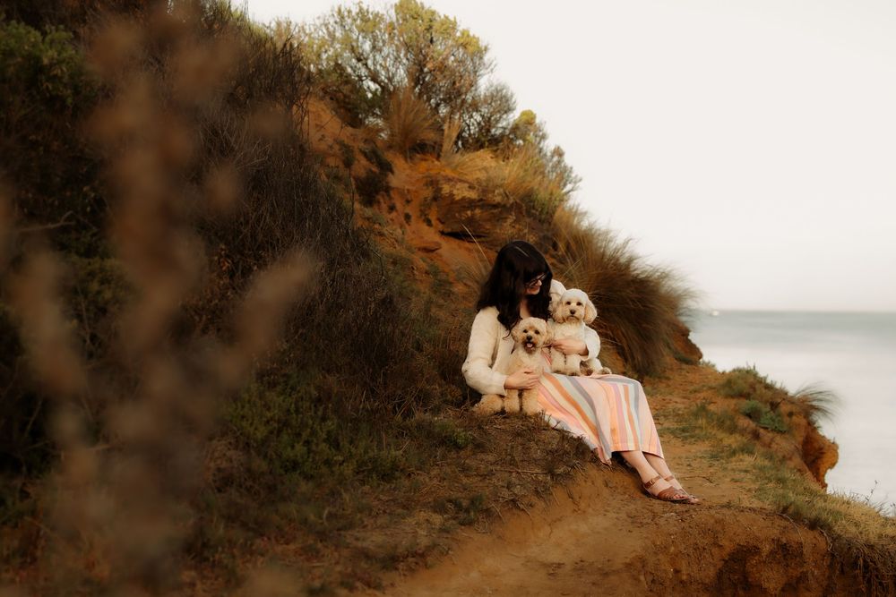 Woman sitting on a grassy coastal cliff holding a fluffy white dog, with the ocean visible in the background.
