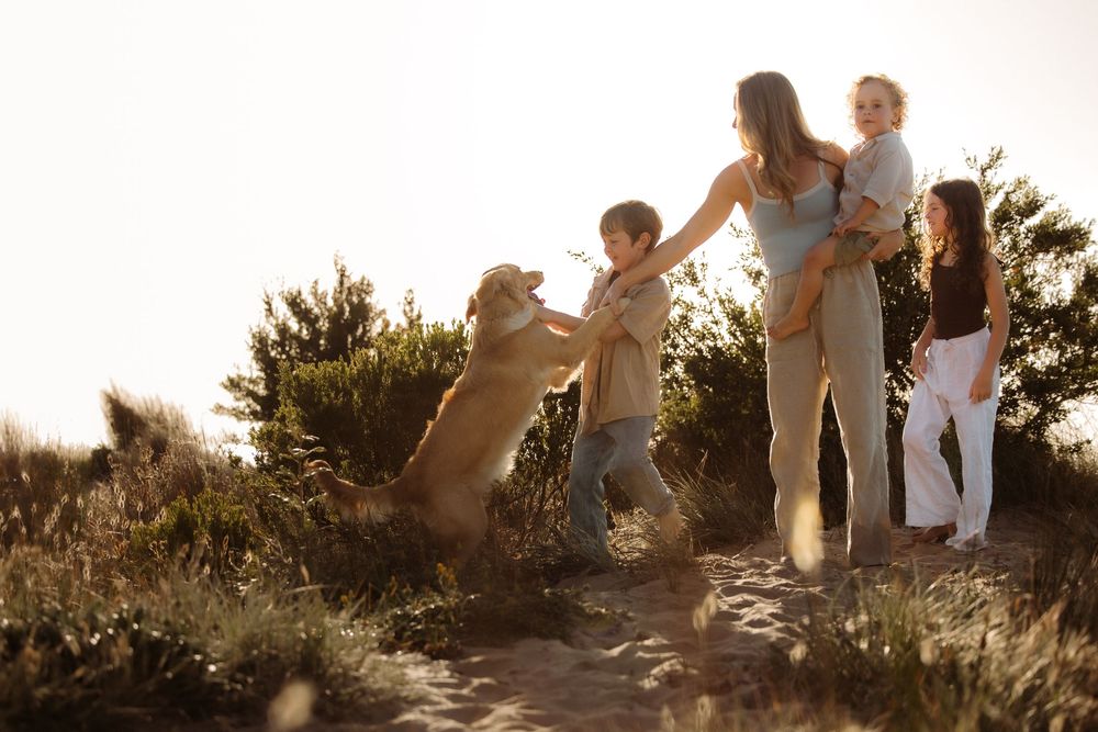Family with golden retriever dog outdoors at sunset, mother holding baby while children play with pet on sandy trail.