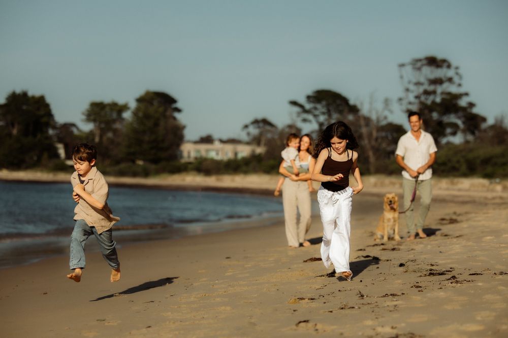 Family with dog running and playing on a sandy beach on a sunny day with trees in the background.