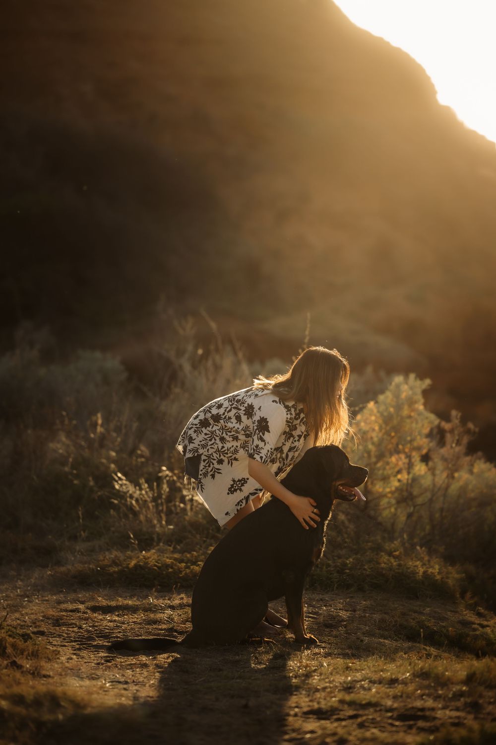 Woman in floral top hugging black dog at golden sunset with mountain backdrop.
