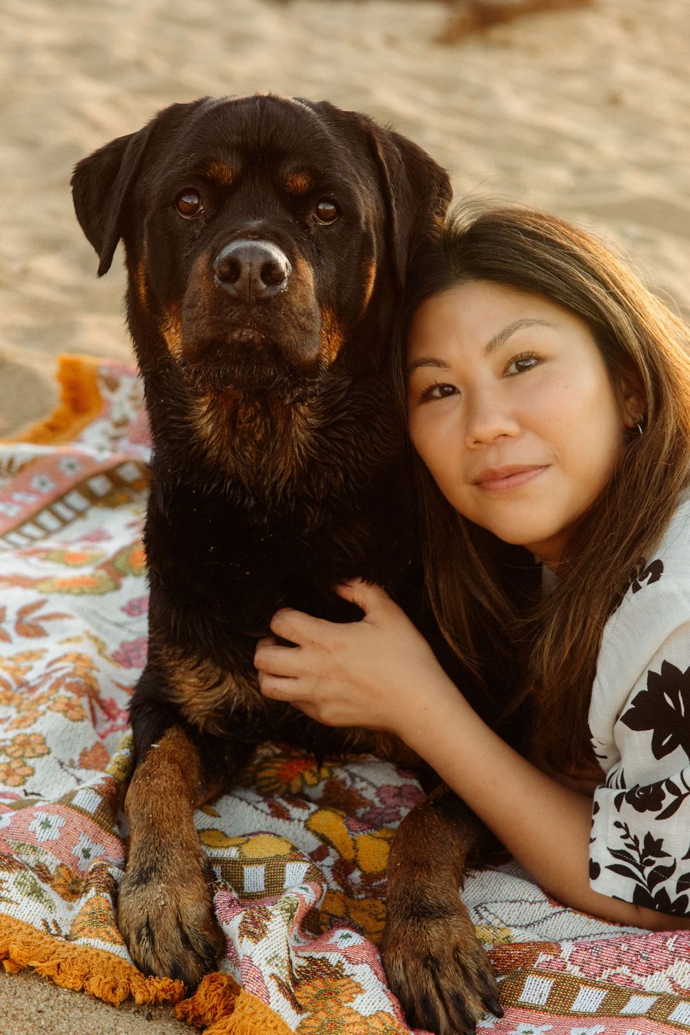 Woman and Rottweiler dog posing together on a colorful beach blanket in golden hour sunlight.