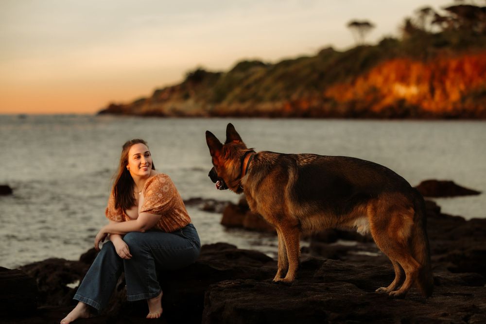 Woman sitting on coastal rocks at sunset with a German Shepherd dog, ocean and golden-lit cliffs in background.