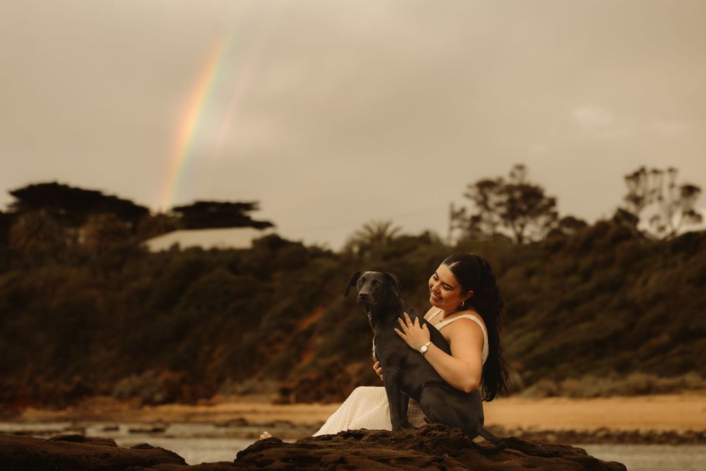 Woman sitting on rocks with black dog, rainbow over ocean beach landscape in background.
