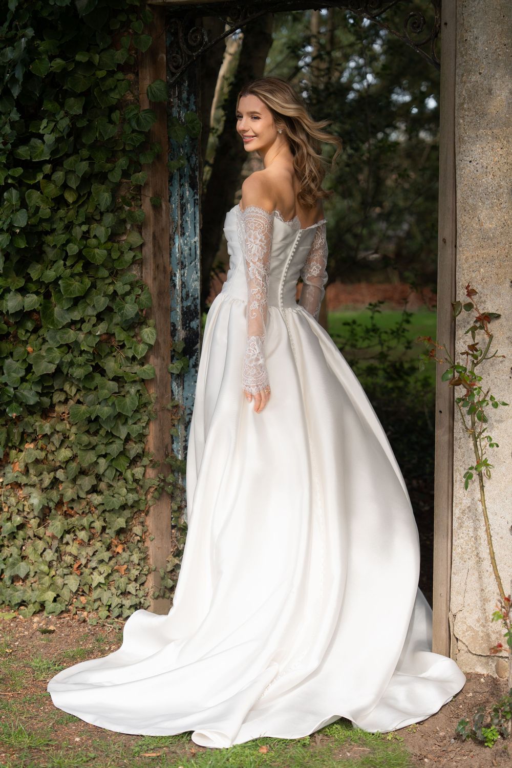 Elegant bride in off-shoulder lace bodice ball gown with long train, posing in rustic garden archway. Cambridge, UK