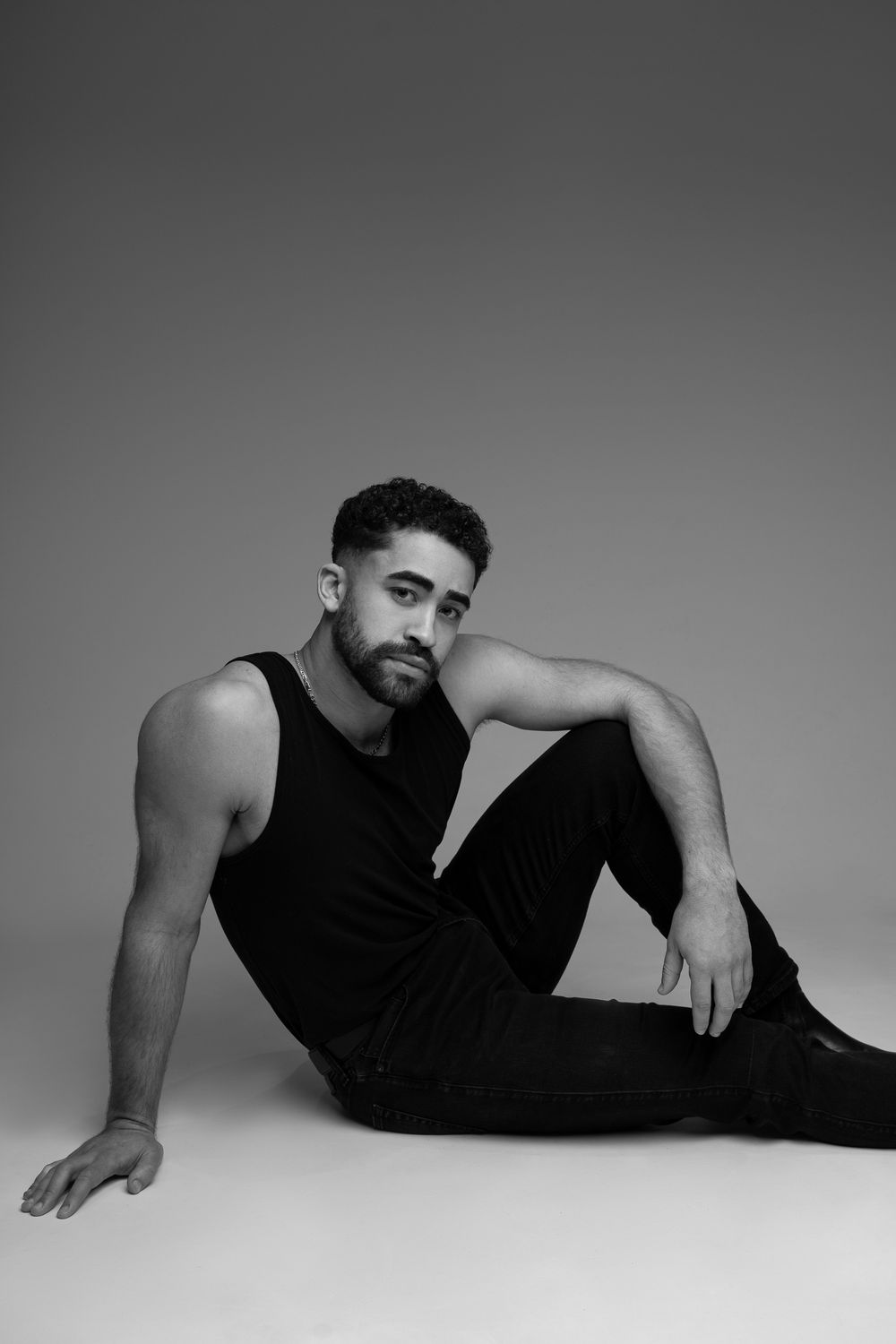Black and white portrait of a man in a tank top and dark pants reclining on floor in artistic pose against neutral background, Cambridge.