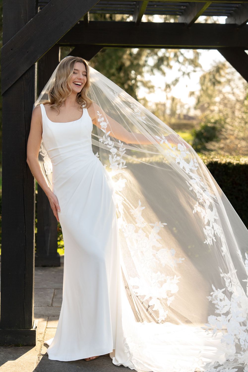 Bride in sleek white gown with floral lace cathedral veil flowing in the breeze, laughing joyfully outdoors, Swynford Manor, Ely