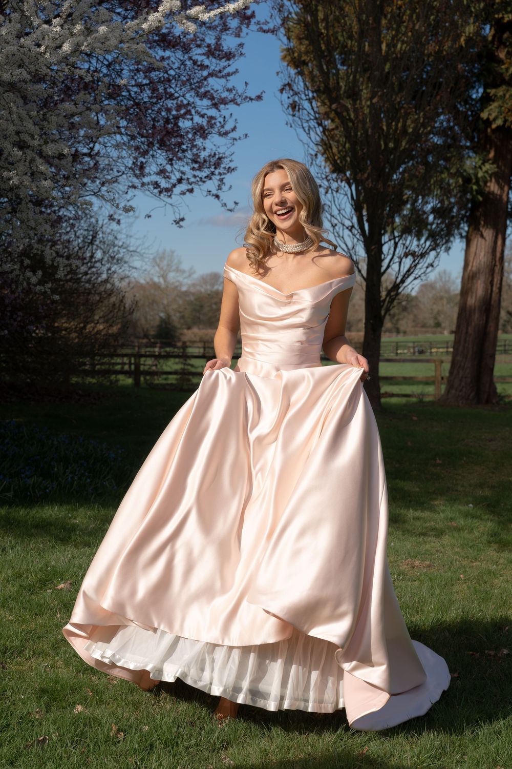 Young woman in blush pink satin off-shoulder ball gown, twirling outdoors in a garden with spring blossoms. Cambridge, UK