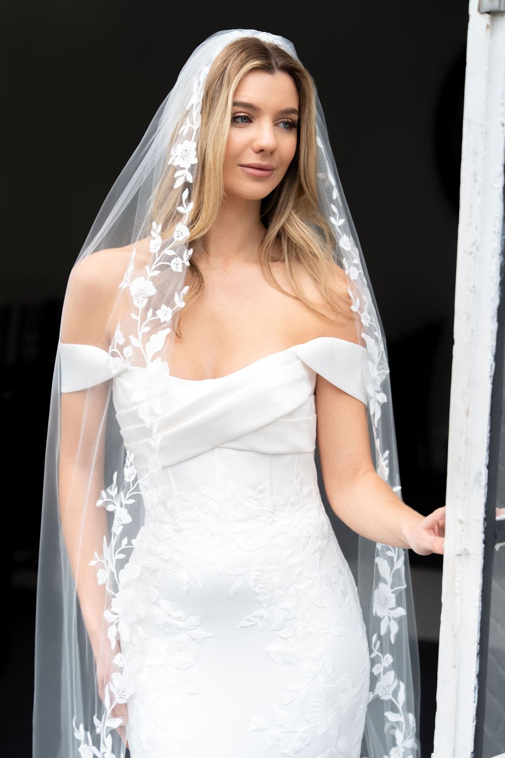 Bride wearing an off-shoulder white lace wedding gown with a floral lace cathedral veil, posing elegantly, at Swynford Manor, Newmarket