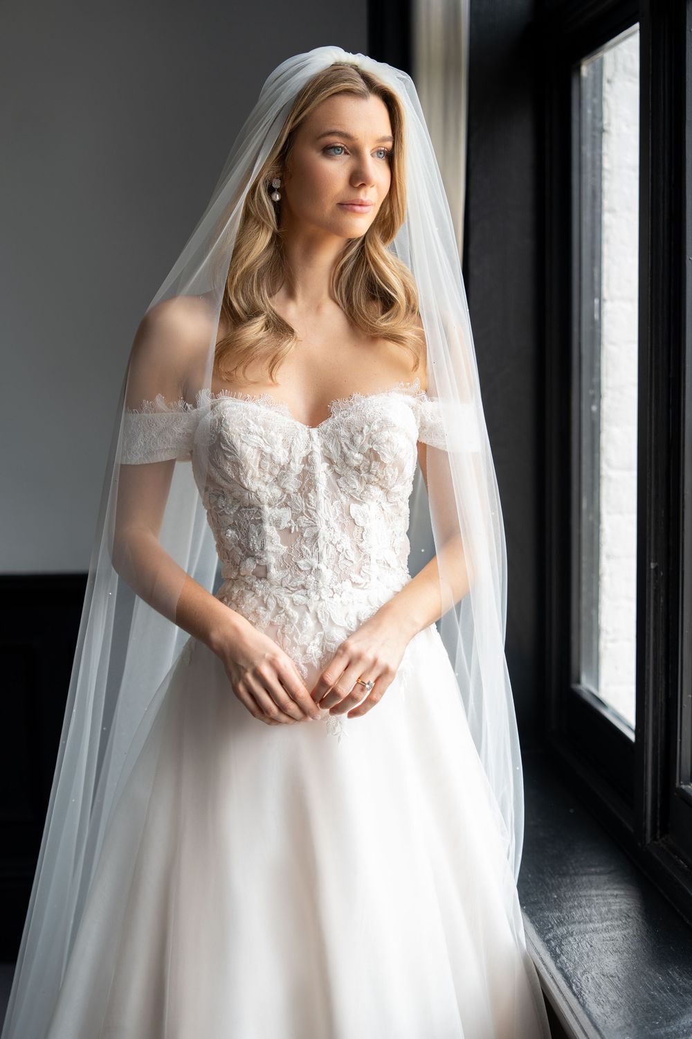 Elegant bride in off-shoulder lace wedding gown with flowing veil stands by the window in soft natural light at Swynford Manor, Newmarket