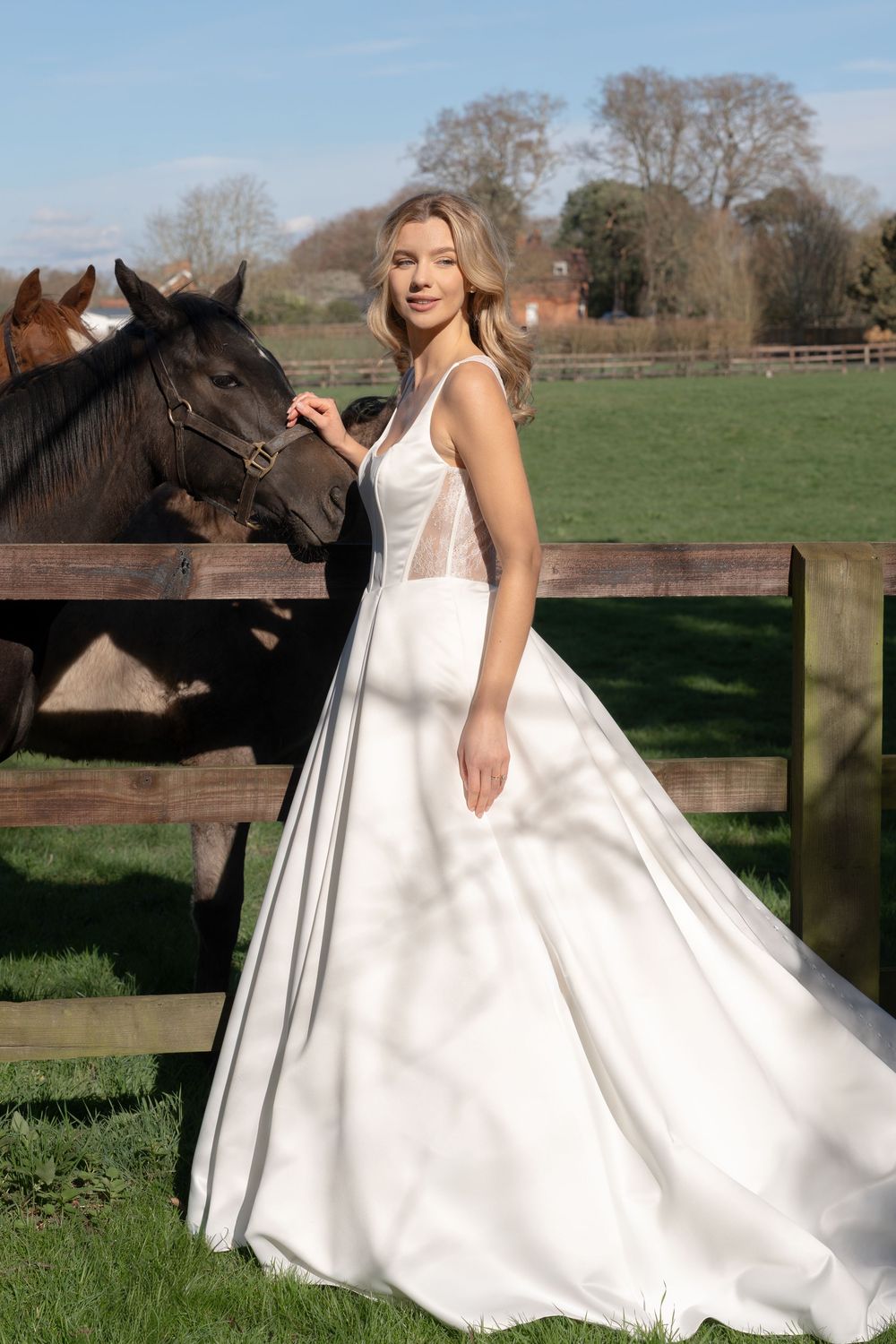 Bride in white ballgown with sheer corset bodice stands beside dark horses at a wooden fence in a green countryside setting. Ely, UK