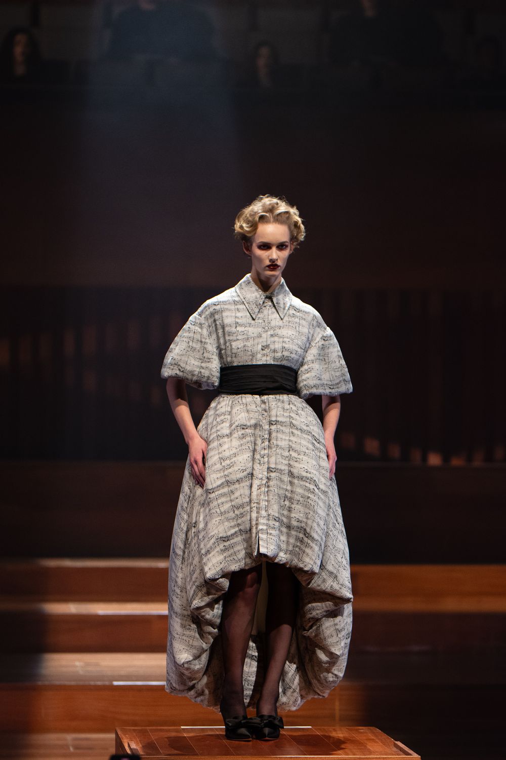 Grey plaid dress with balloon sleeves and black waist belt presented on wooden runway.