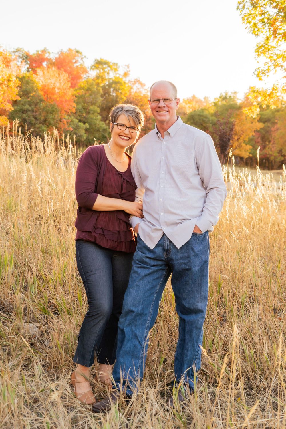A couple stands together in a sun-lit autumn field with golden grass and fall foliage.