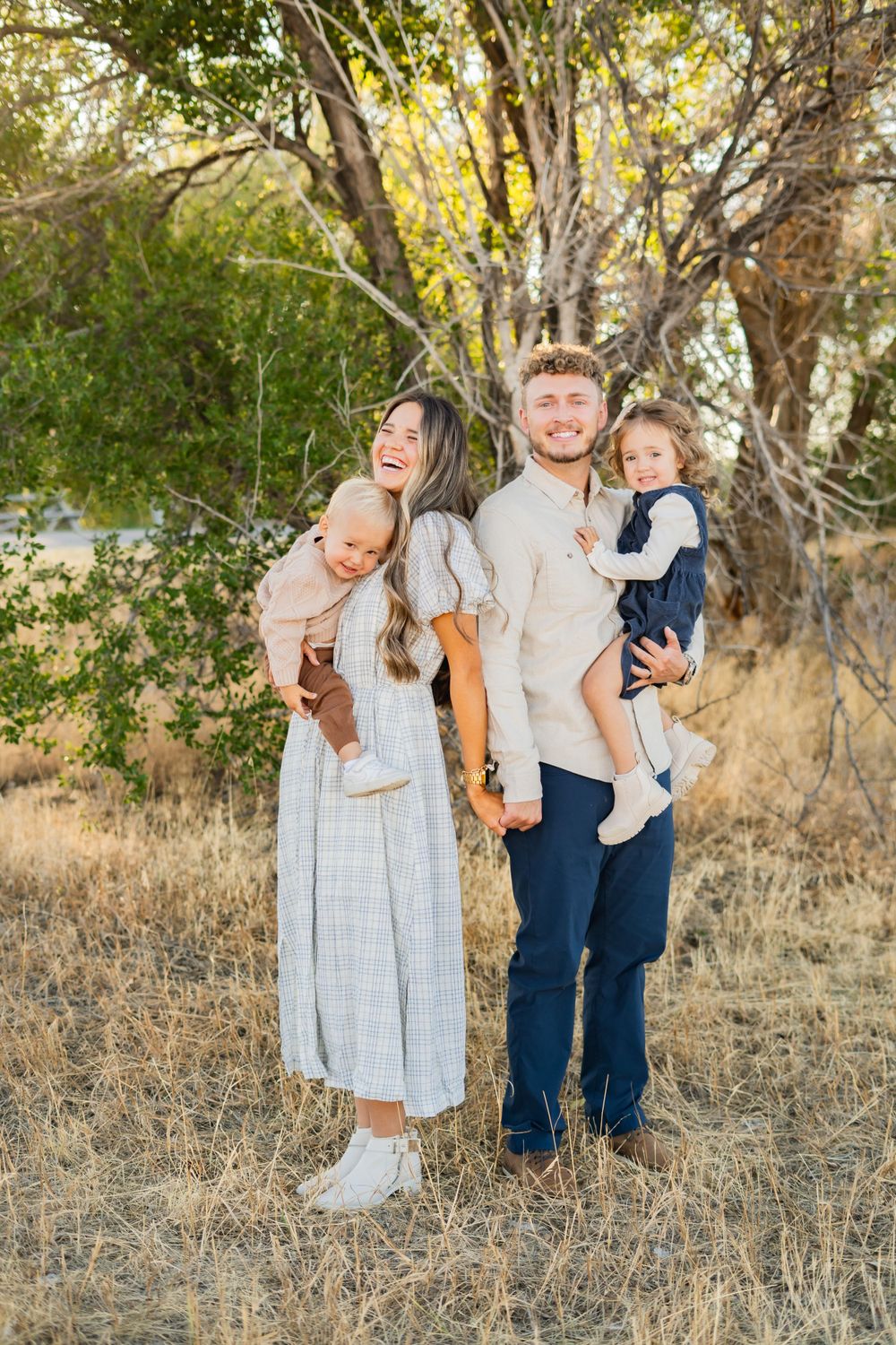 A young family stands together in a natural setting with trees and dry grass.