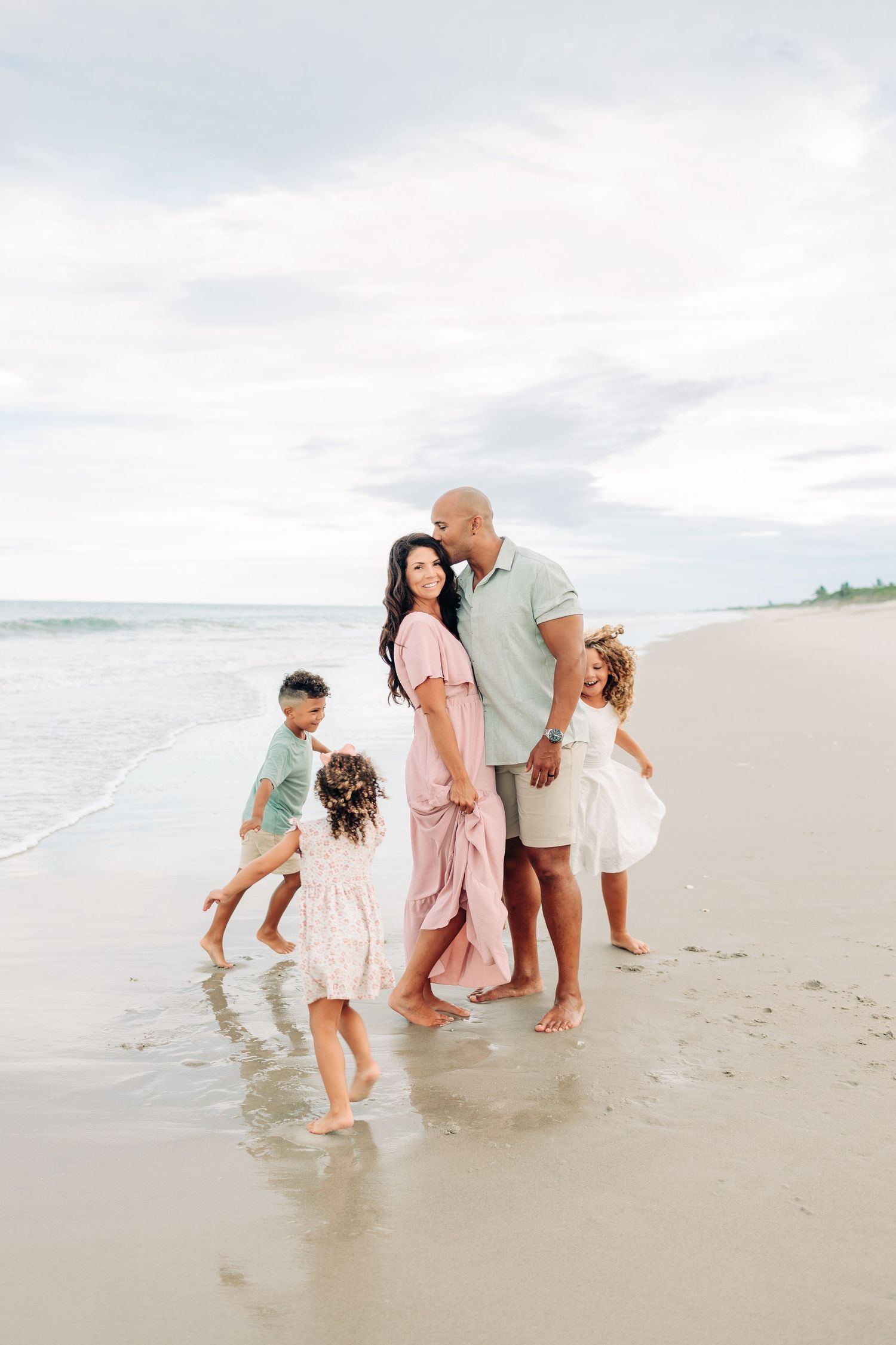 Candid moment of dad kissing mom while their children run around them during a sunset beach family photo session in Orlando