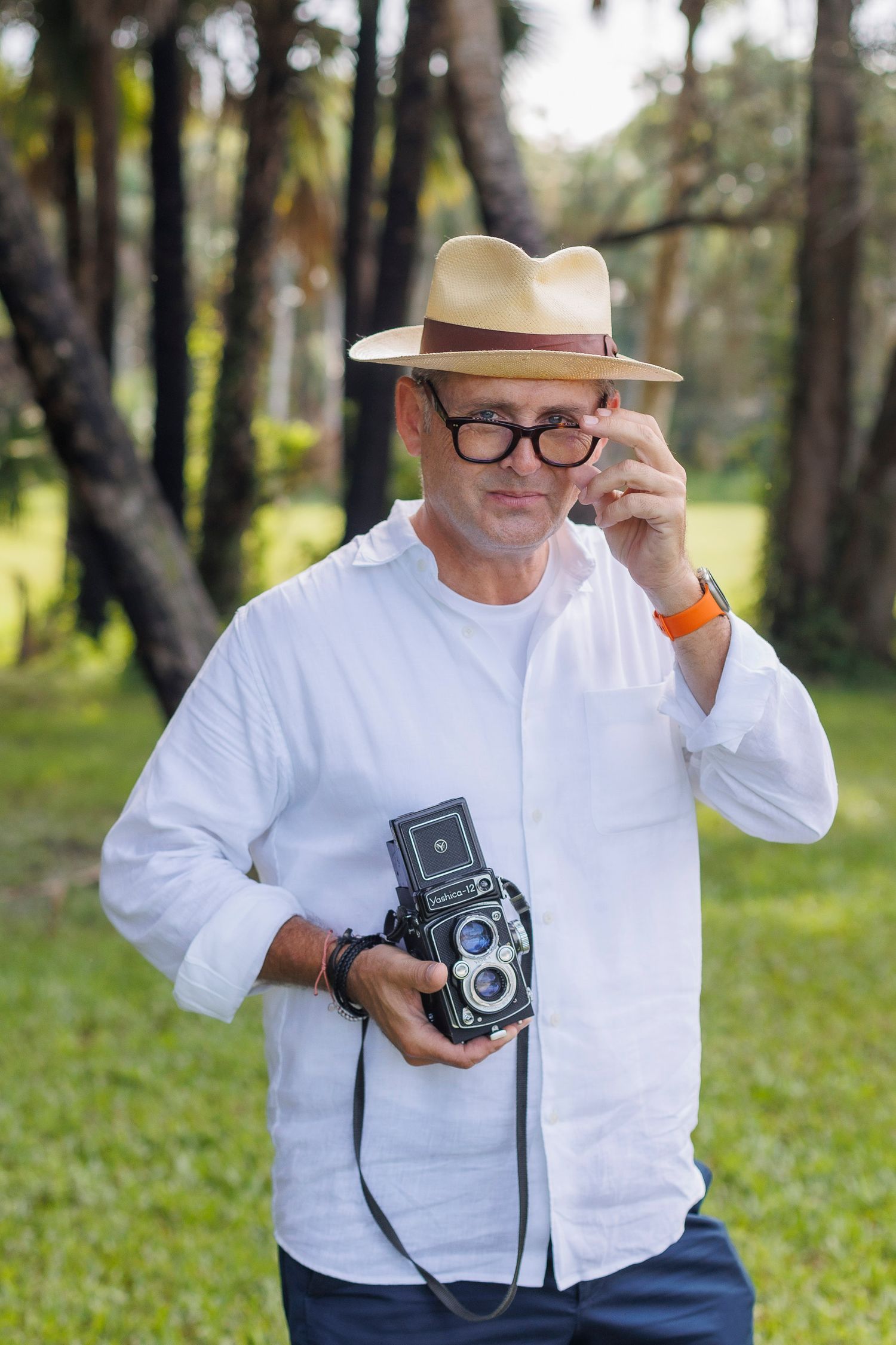 Photographer in white shirt and fedora holds vintage twin-lens camera outdoors.