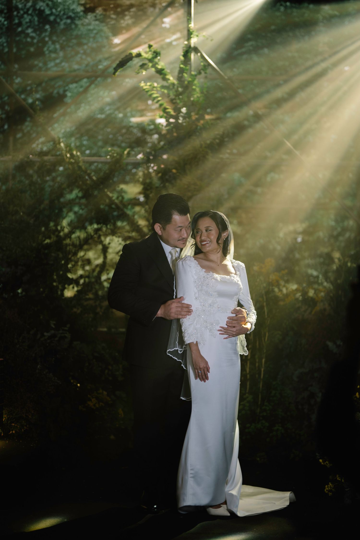 Romantic wedding couple embracing in a sunlit forest with beautiful rays of light shining through trees.