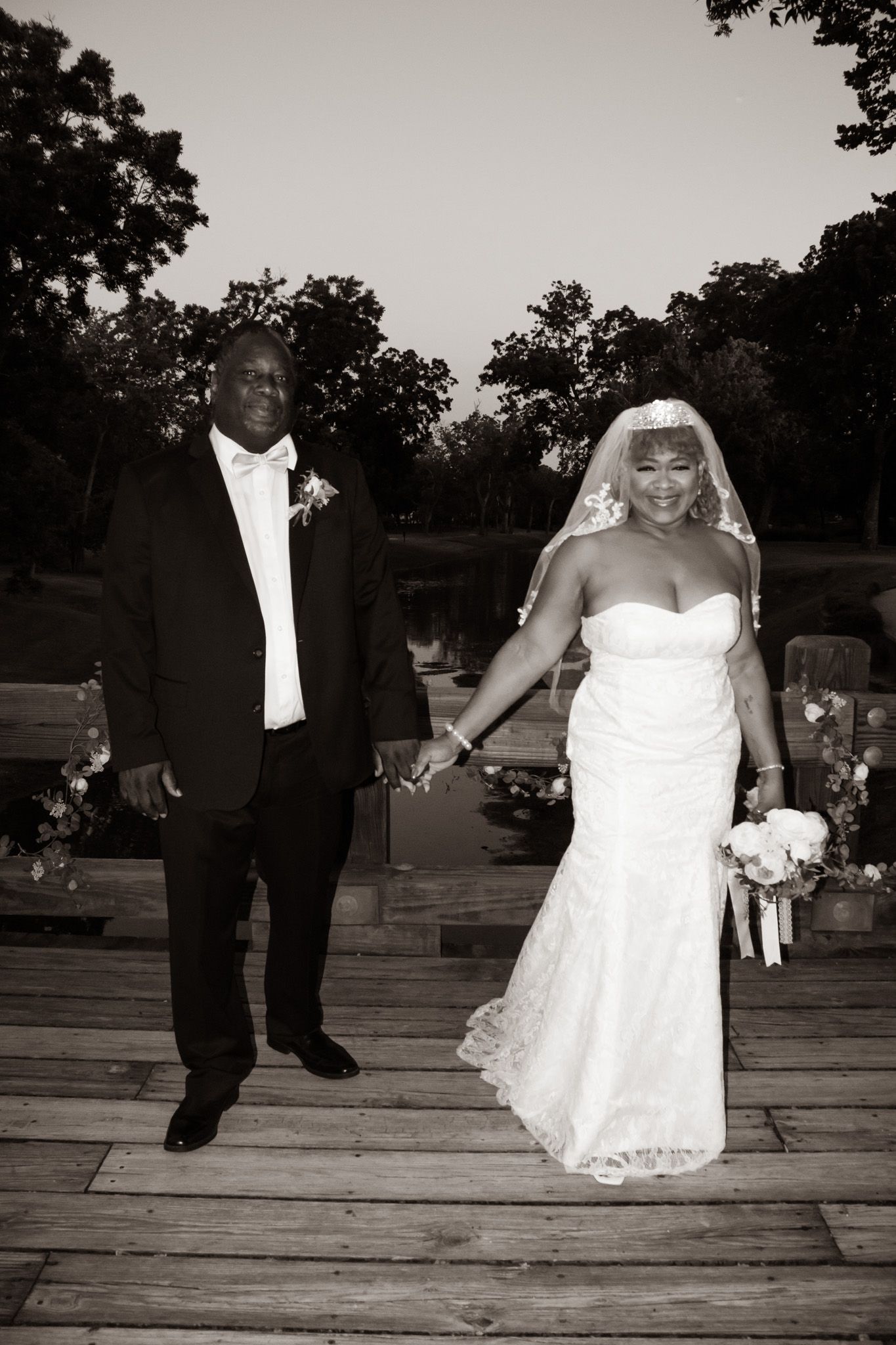 Black and white photo of a bride and groom walking hand in hand on a wooden outdoor aisle.