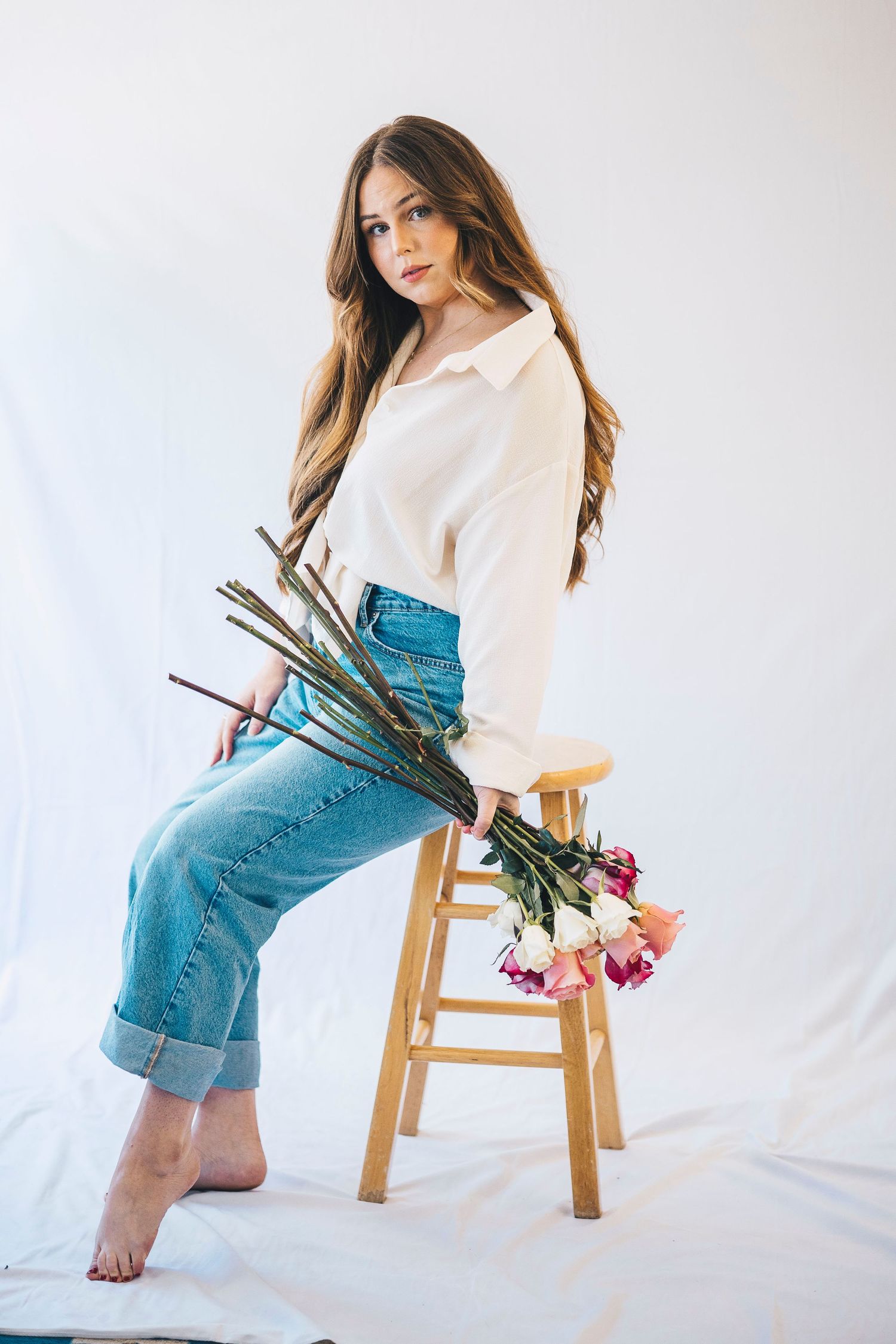 Young woman in white blouse and jeans sitting on wooden stool holding colorful roses.