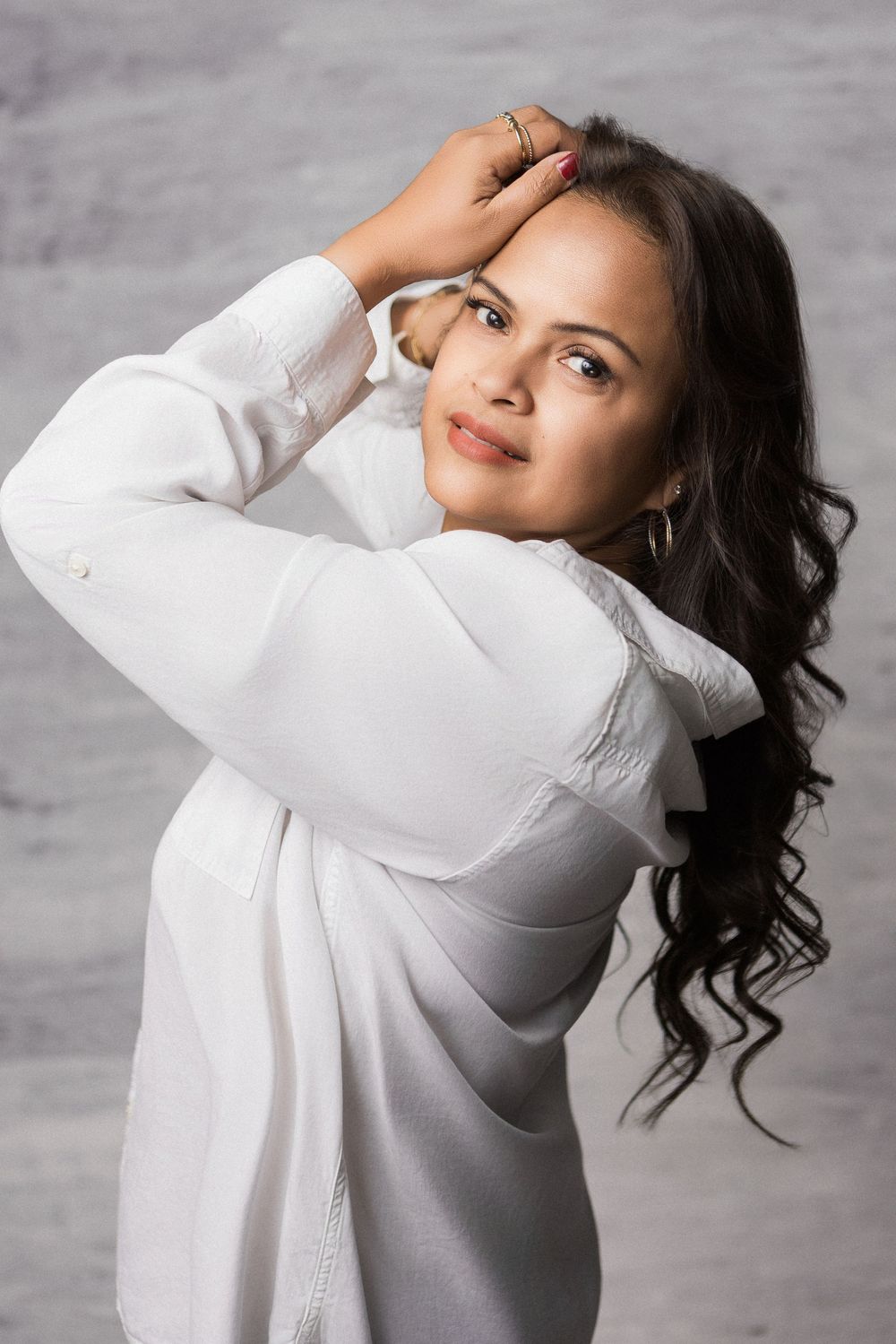 Young woman with long dark wavy hair poses in white shirt, hands behind head, smiling softly against gray background.