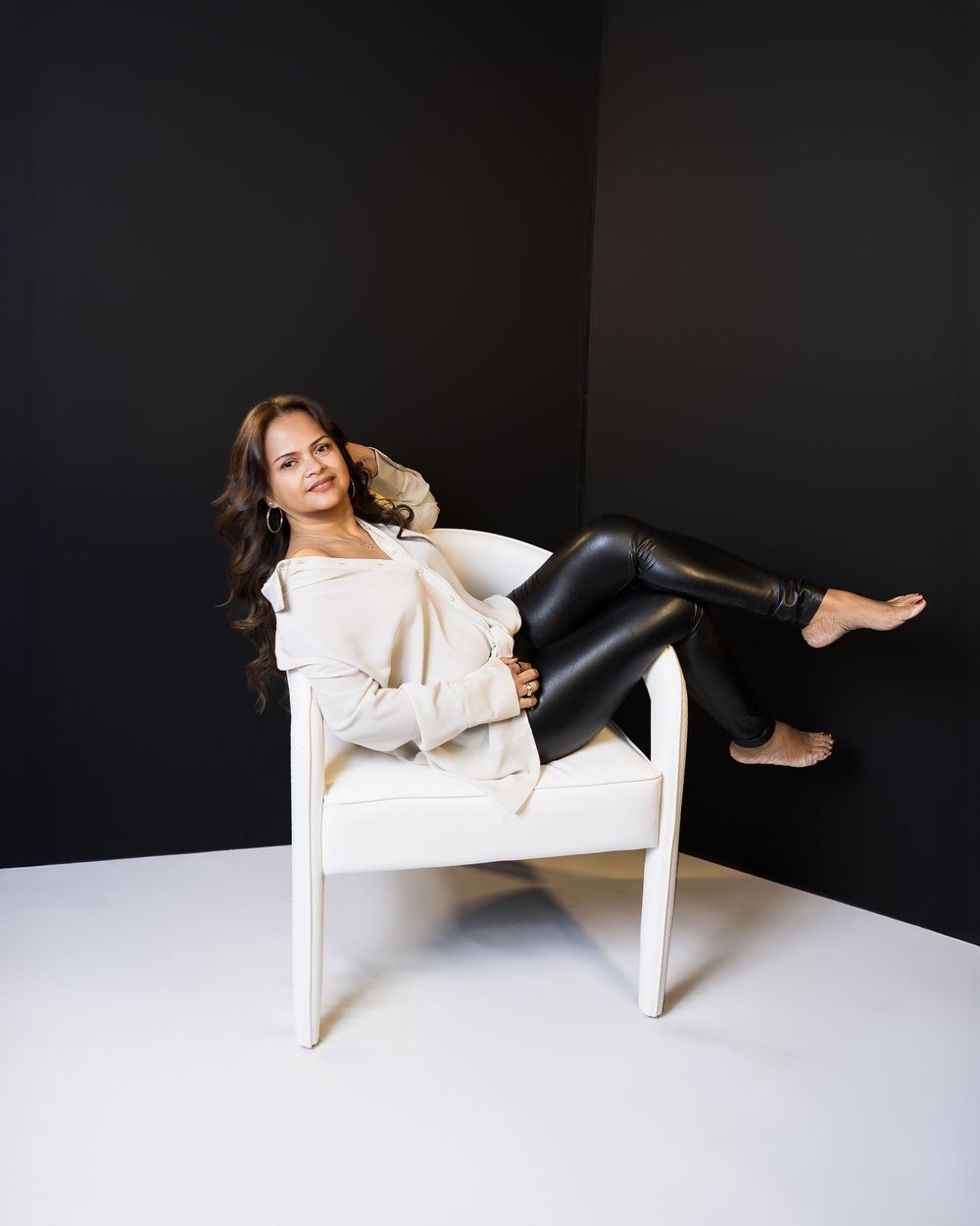 Woman in white top and black leather leggings poses relaxed on a white chair against a dark wall.