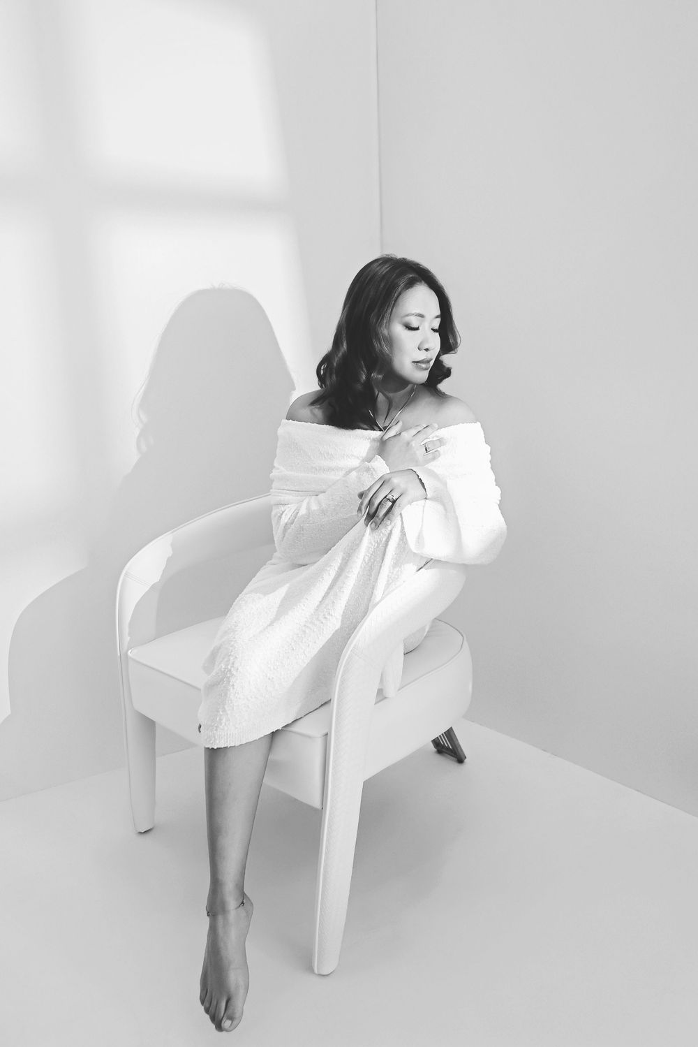Black and white photo of a woman in an off-shoulder white dress sitting gracefully on a white chair in a bright studio.