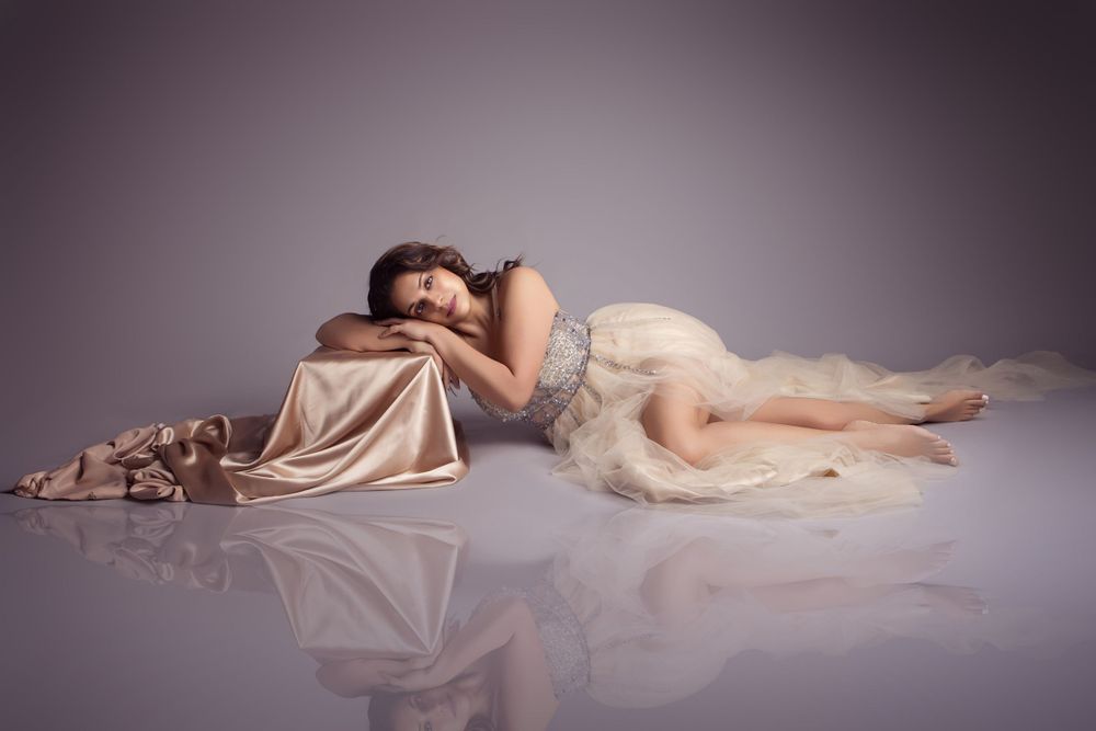 A graceful figure reclines in a dreamy pose wearing an elegant cream gown on a reflective surface.