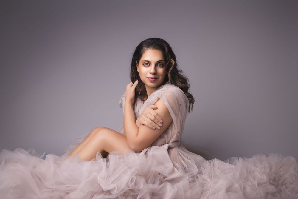 A figure sits elegantly on the floor surrounded by layers of flowing blush pink tulle fabric in a dreamy studio setting.