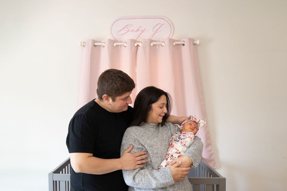 Parents cradle their newborn baby in front of a pink curtain in a nursery setting.