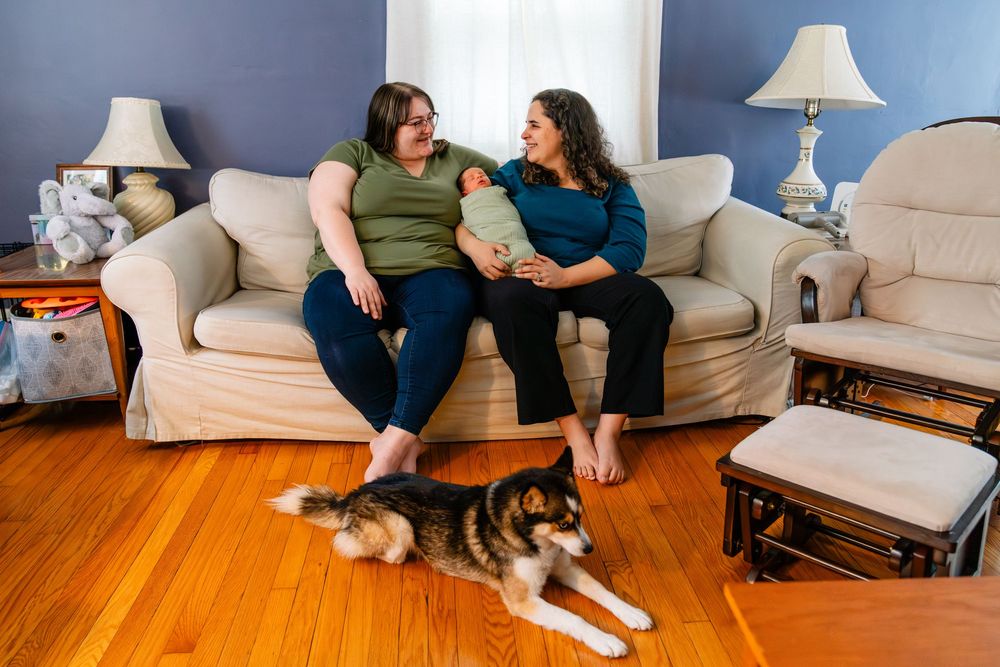 A couple relaxes on a beige couch in their living room with a dog laying on the hardwood floor.