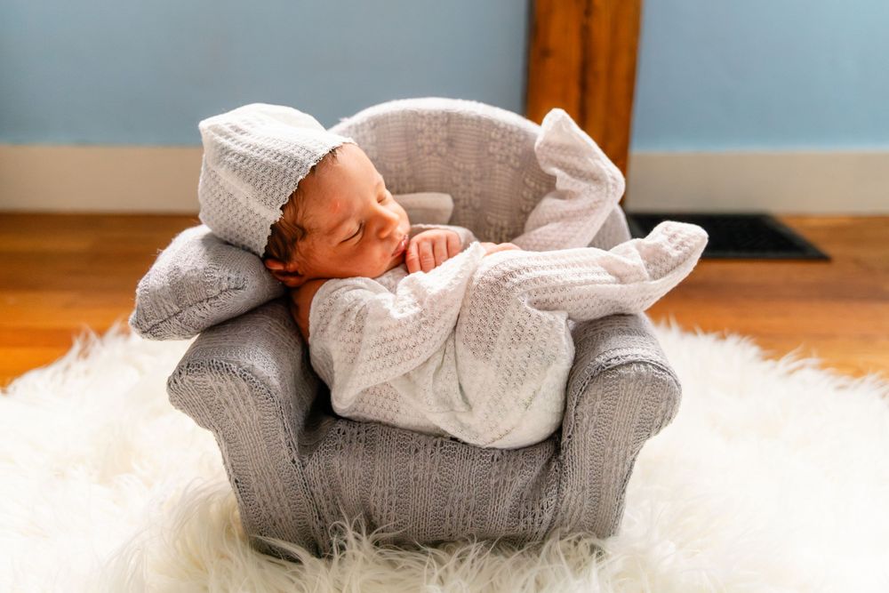 A tiny newborn sleeps contentedly in a miniature gray armchair on a white fluffy rug.
