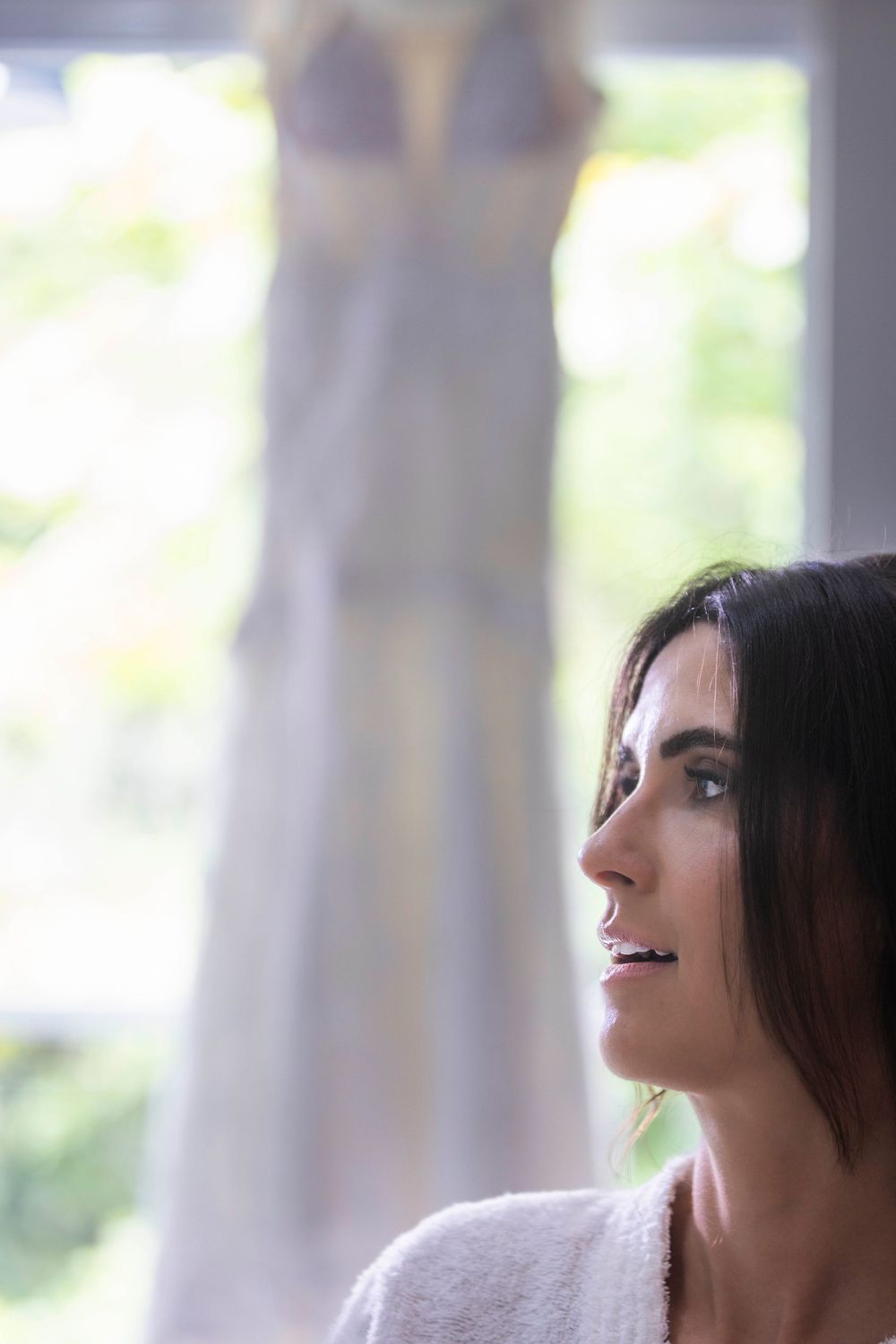 Bride in white robe gazes thoughtfully with her wedding dress hanging in a sunlit window behind her.