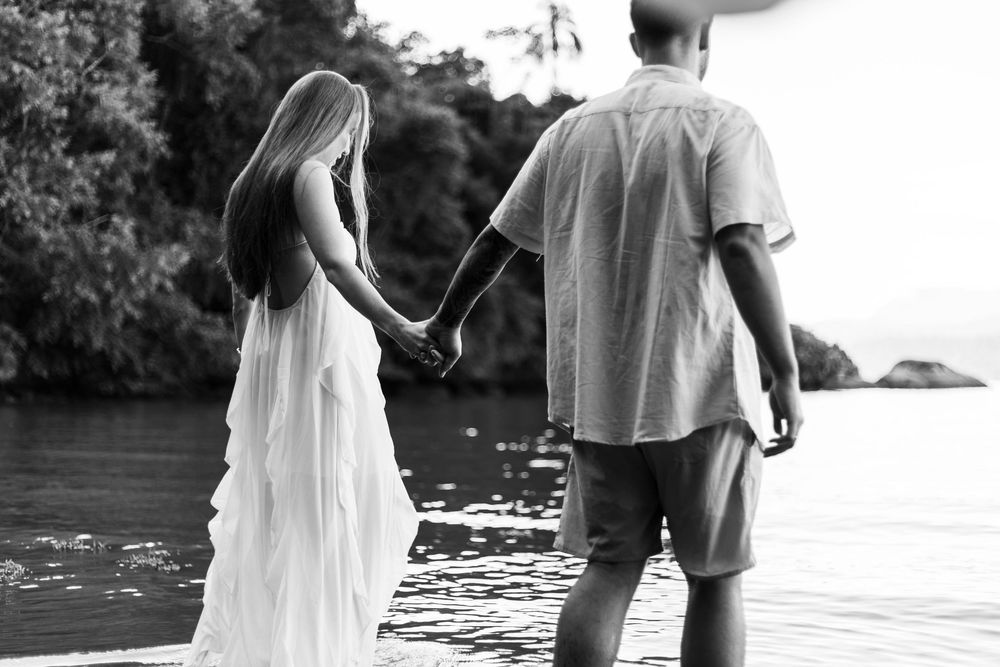 Black and white photo of a couple holding hands on a dock by a serene lake surrounded by lush trees.