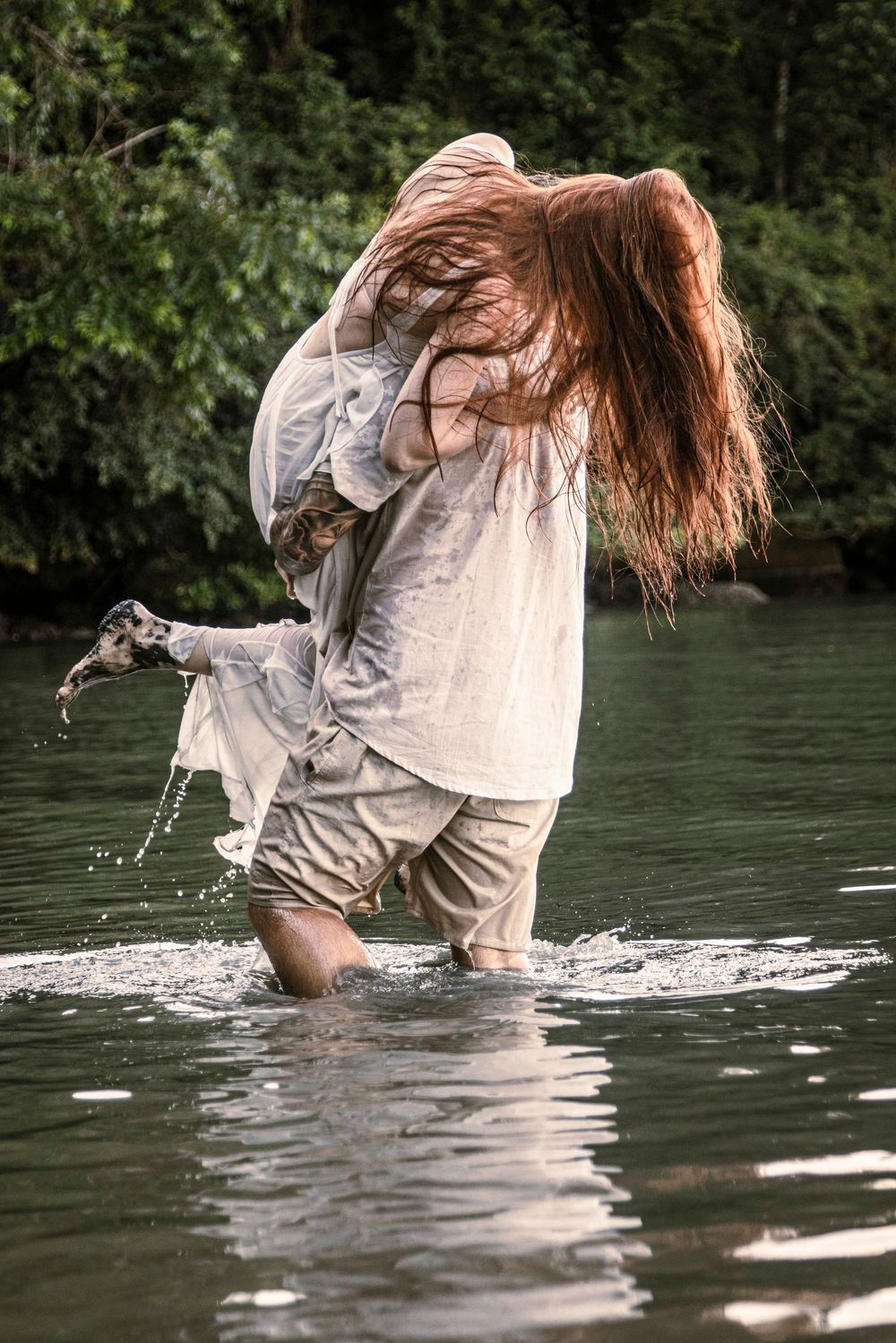 Person with long red hair wading through a river in wet linen clothing, surrounded by lush green trees.