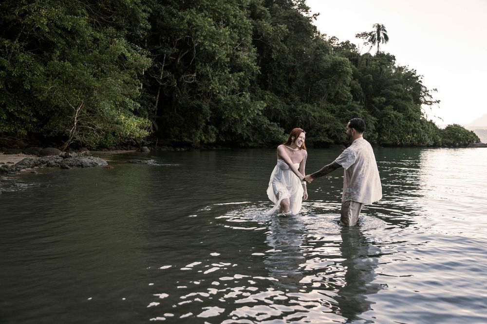 Couple in white attire wading through calm tropical waters surrounded by lush green trees.